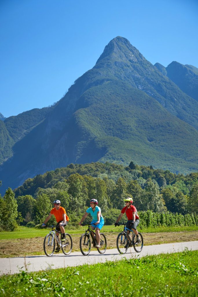 3 turistas de bicicleta no vale do rio Soca com os Alpes ao fundo, na Eslovênia