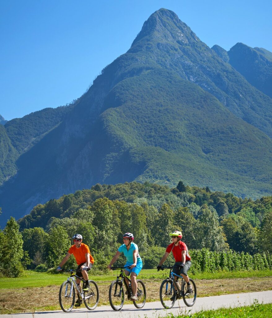 3 turistas de bicicleta no vale do rio Soca com os Alpes ao fundo, na Eslovênia